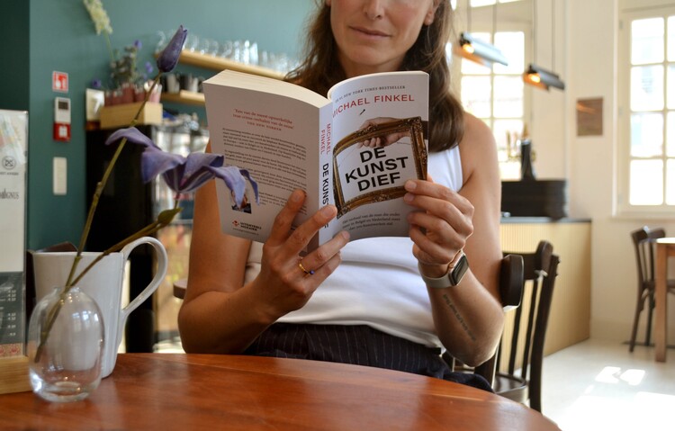 Een persoon zit in een zonnige koffiebar en leest het boek met de titel De Kunstdief.