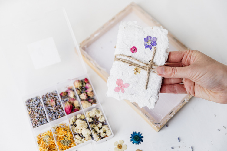Een hand houdt een bundel handgeschept papier omhoog. Op de tafel er onder liggen een schepraam en enkele bakjes vol gedroogde bloemen.