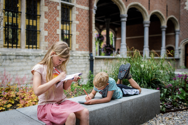 Deux enfants sont assis sur un banc dans le jardin du musée. Ils écrivent ou dessinent dans un livret.