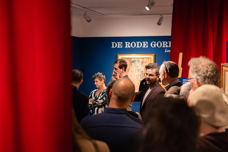 Een groep mensen staat in een museumzaal met blauwe muren en rode gordijnen.