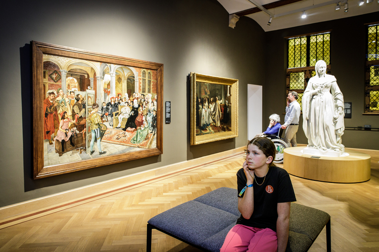 Een kind zit op een grijze bank in een museumzaal. Op de achtergrond is het schilderij van Willem Geets, Poppenspel aan het Hof van Margareta van Oostenrijk, te zien.