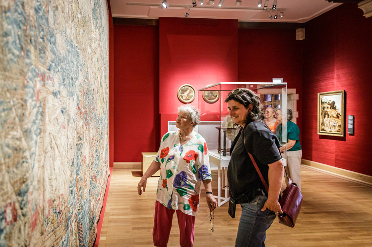 Twee mensen wandelen in een museumzaal en kijken naar een monumentaal wandtapijt.